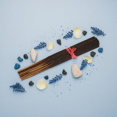 China Rain Incense Sticks with a red ribbon surrounded by stones, flowers, and candles on a light blue background